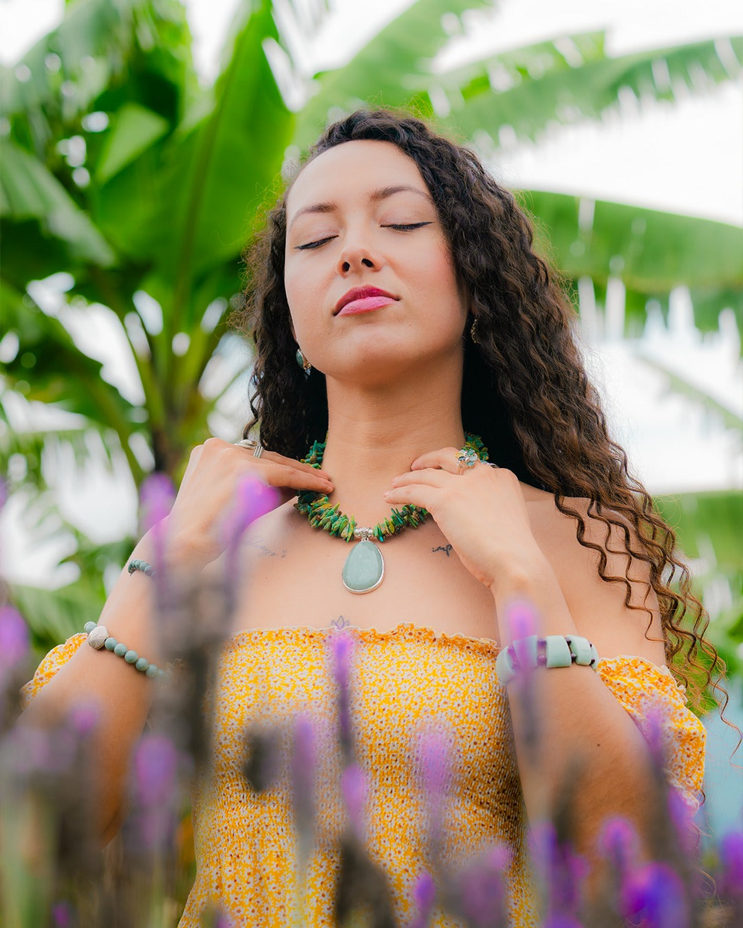 Woman in a yellow dress with a green necklace standing among purple flowers and green foliage.